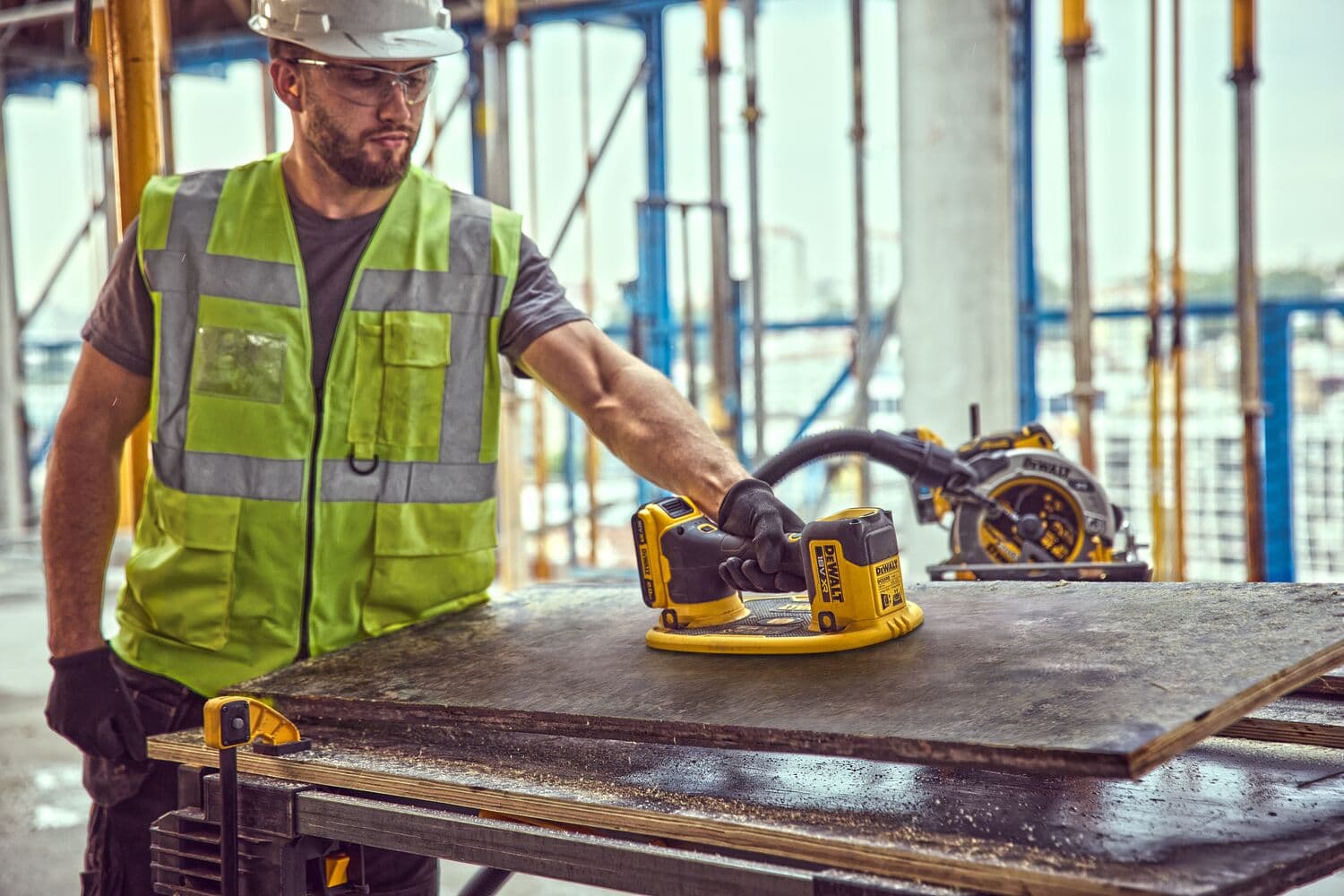 A person wearing a safety vest and gloves uses a DEWALT DCE590 power tool to work on a piece of wood at a construction site. Other DEWALT tools are visible in the background.