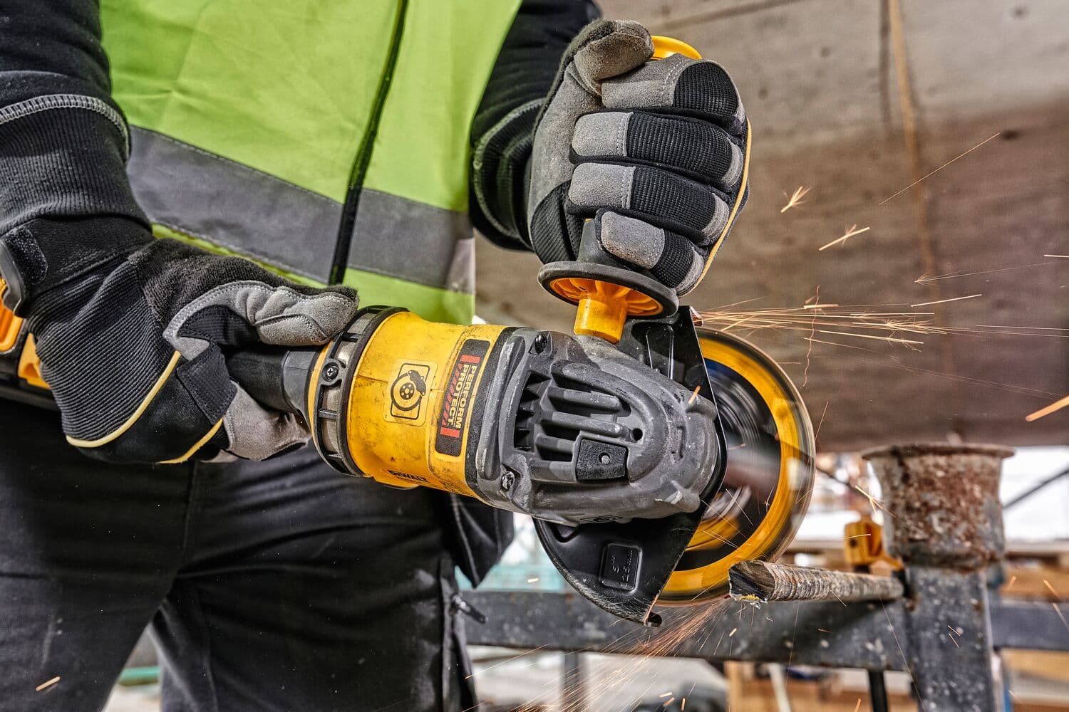 A person wearing safety gloves and a high-visibility vest is using a yellow angle grinder to cut through a metal rod, creating sparks in a construction environment.